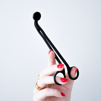 A hand with red nail polish holds a black wick trimmer for candles against a plain light gray background.