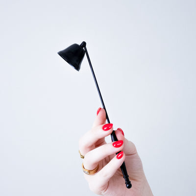 A hand with red nail polish holds a small black metal candle snuffer against a plain light gray background.