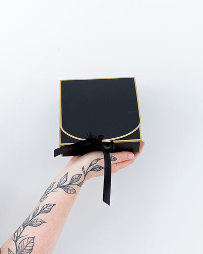 A hand with leaf tattoos holds a black gift box with gold trim and a black ribbon bow against a plain white background.