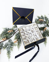 An open advent calendar black gift box with gold trim and a black ribbon, filled with wax melts, styled with green pine branches and gold berries on a white background.